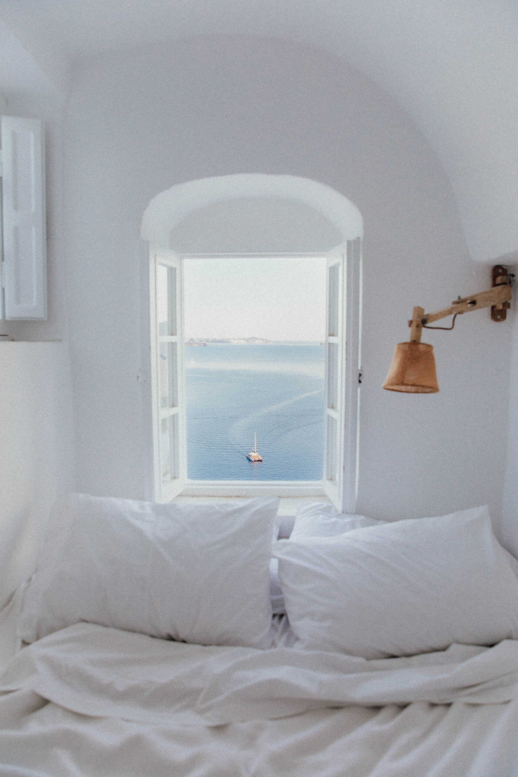 A cozy Santorini bedroom with white linens, natural light, and a window view of the Aegean Sea